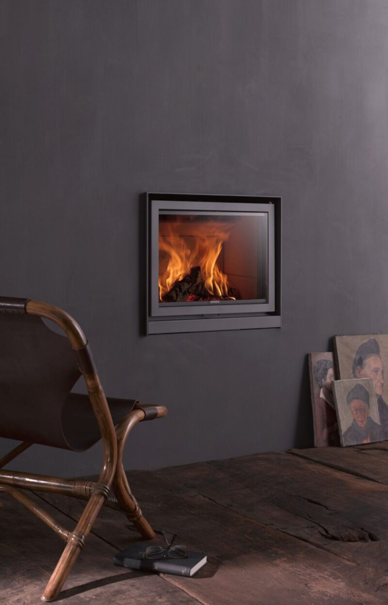 A modern room featuring a dark gray wall with a mounted electric fireplace displaying lively flames. a sleek wooden chair and a rustic wooden table are in the foreground, complementing the minimalist decor.