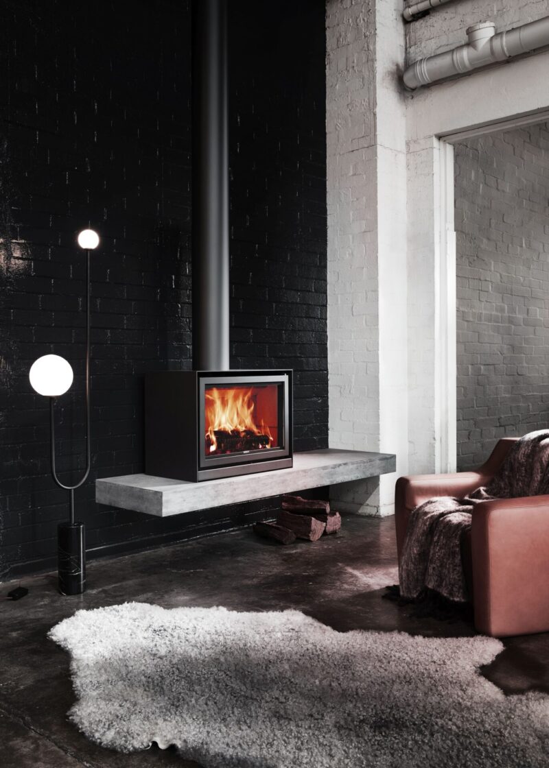 A modern, stylish living room with dark brick walls featuring a sleek, freestanding fireplace on a concrete platform, a plush chair, a floor lamp, and a textured rug.