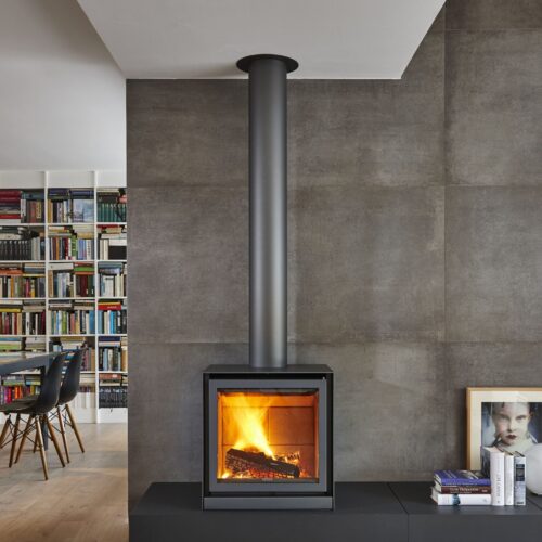 Modern living room with a sleek, wall-mounted fireplace, extended metal flue, and a full bookshelf in the background. concrete walls add an industrial touch, with a small seating area to the side.