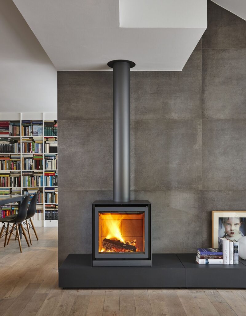 Modern living room with a sleek, wall-mounted fireplace, extended metal flue, and a full bookshelf in the background. concrete walls add an industrial touch, with a small seating area to the side.