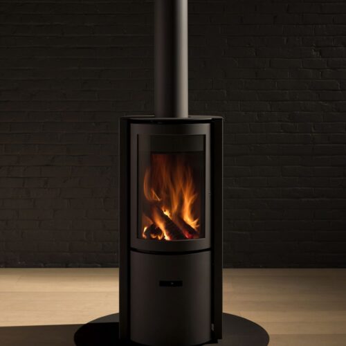 A modern, black wood-burning stove with a visible flame, set against a dark brick wall, and resting on a wooden floor under a centered metallic chimney.