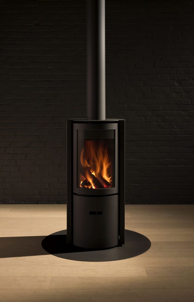 A modern, black wood-burning stove with a visible flame, set against a dark brick wall, and resting on a wooden floor under a centered metallic chimney.