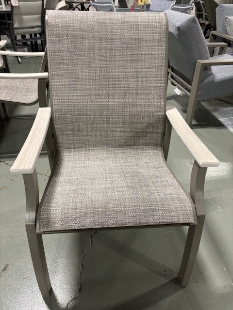 A St. Catherine Sling dining chair with textured light beige upholstery and gray wooden arms and legs, displayed in a furniture showroom setting.