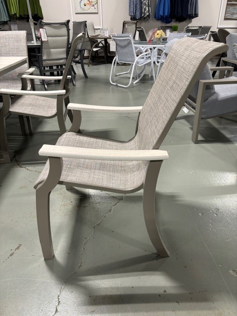 A beige, upholstered St. Catherine Sling outdoor reclining chair with armrests, displayed in a store among other patio furniture. The chair is placed on a green concrete floor.