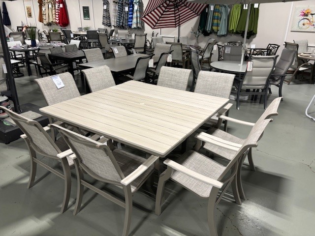 A furniture showroom displaying various outdoor dining sets including St. Catherine Sling chairs and tables in different styles and materials, arranged neatly on a grey floor.