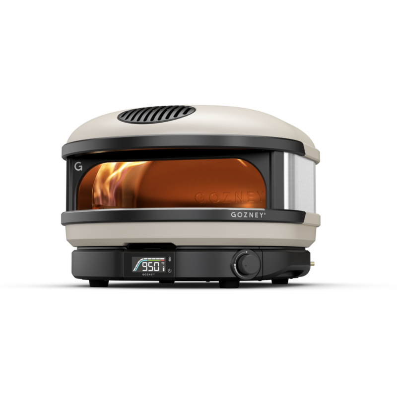 A gozney portable pizza oven with a visible flame through its front glass, digital temperature display reading 950, set against a plain background.
