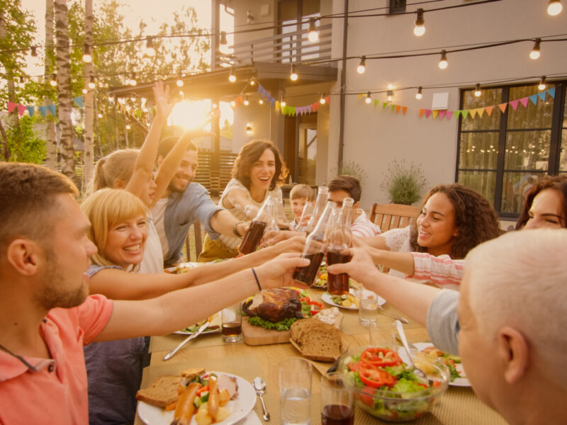 A broad view of a group of smiling people standing and leaning across the outdoor dining table to clink glasses; outdoor string lights and the setting sun offer additional illumination