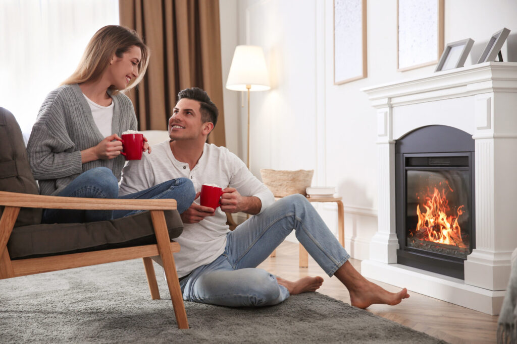 A happy couple gazes at each other while holding mugs and sitting in front of a fire with a white mantel and surround