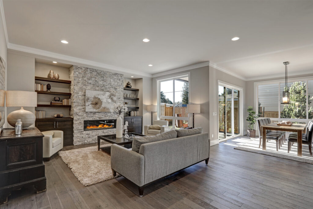 A modern fireplace in a stone wall serves as the focal point of an open concept living room with wood floors, a gray couch, and plenty of natural light