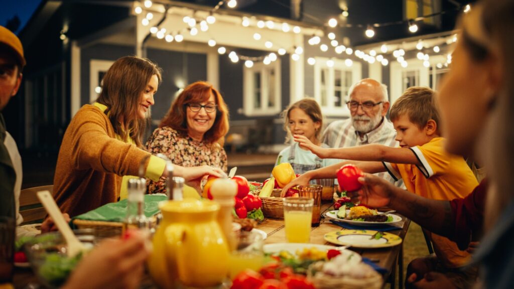 A multigenerational group sits around an outdoor dining table, smiling and reaching for food, with lights hung above with the exterior of the home in the background