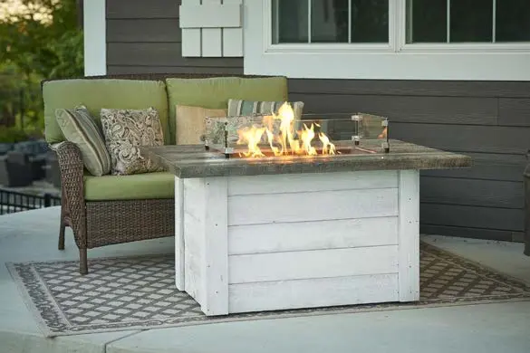 A lit outdoor fire table with a white-washed wood base on a patio with a rattan settee with green cushions and multi-colored throw pillows