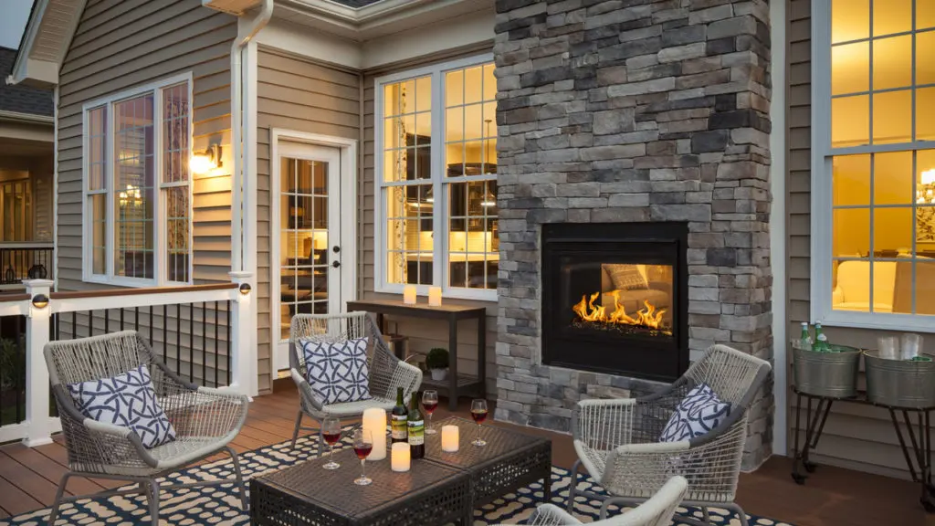 A lit, modern outdoor fireplace in a stone surround offers indoor-outdoor ambiance for a patio with several chairs and a cocktail table