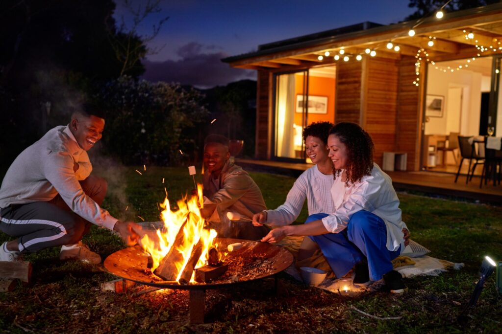 Four smiling people sit around an outdoor firepit with an illuminated home and the evening sky in the background