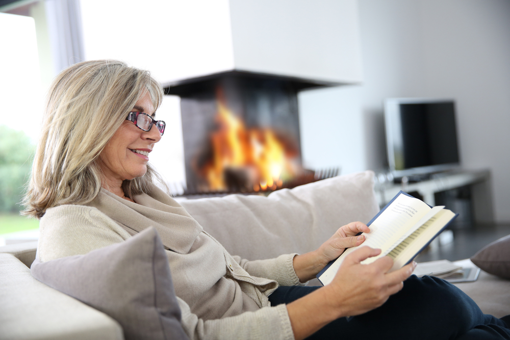 A woman in a tan sweater and wearing glasses reads in a brightly lit living room while relaxing on a sofa near a lit fire