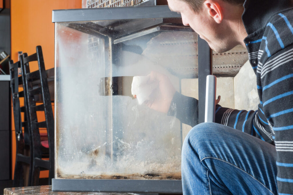 A homeowner in a blue striped shirt and jeans wipes white residue while cleaning fireplace glass  